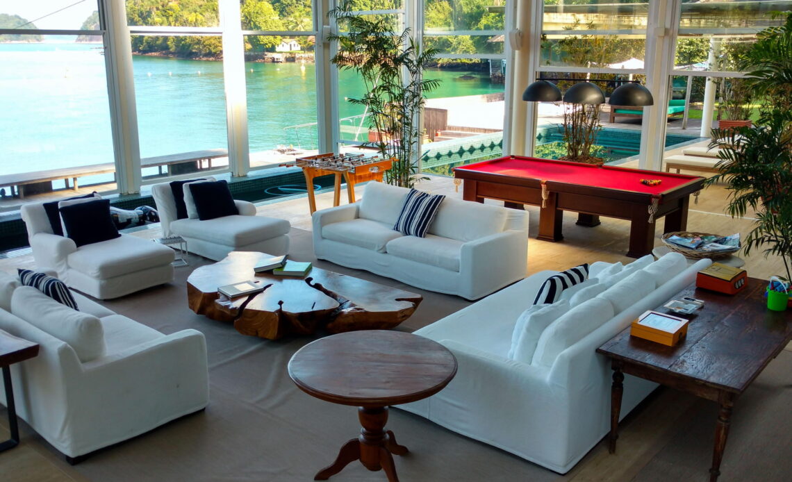Morar na Praia | Foto da sala de estar com sofas brancos e destaque para mesa de sinuca vermelha, ao fundo a vista remete ao mar de angra.