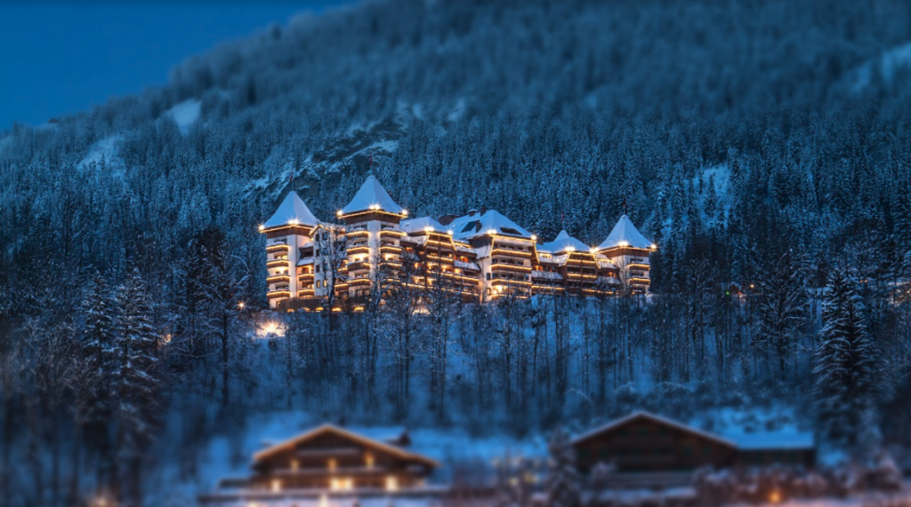 The Alpina Gstaad| Foto panoramica do hotel mostrando ele e seus arredores, em tons de azul e branco neve todo iluminado