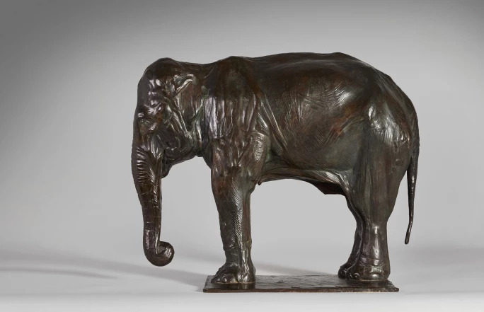 Foto da peça escultural realística de um elefante asiático, sobre uma base reta e estreita do mesmo material e cor da escultura. Peça do escultor Rembrandt Bugatti.