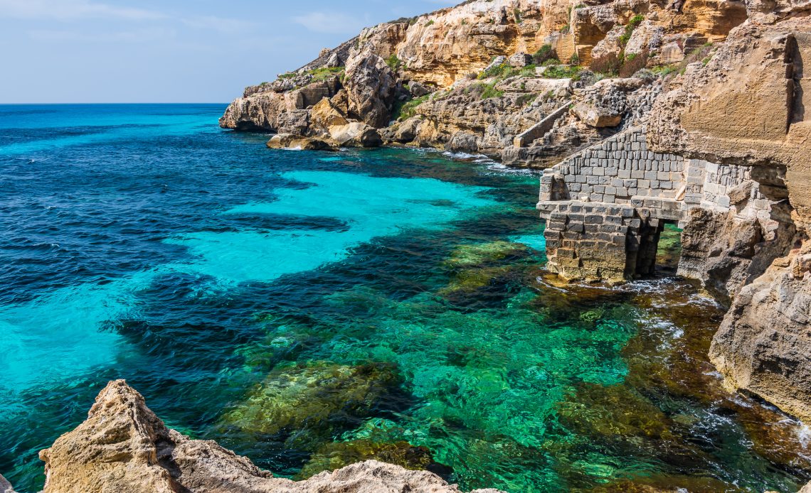 Foto de falésias da ilha Favignana, na costa oeste da Sicília. É possível ver costões rochosos à direita, com ruínas de blocos de pedra, circundando o mar azul e transparente. A clareza da água permite vislumbrar pedras, corais e algas submersos.