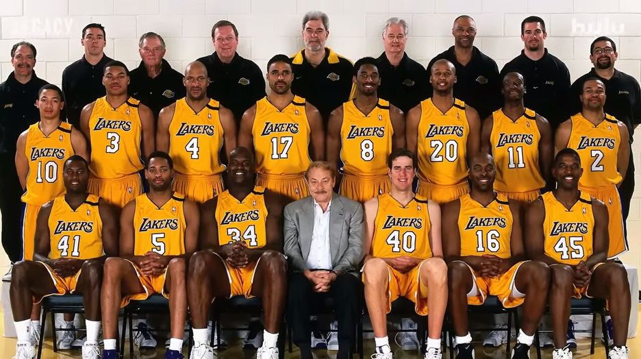 Time de basquete reunido vestidos com o uniforme laranja dos Lakers junto ao dono e a comissão técnica