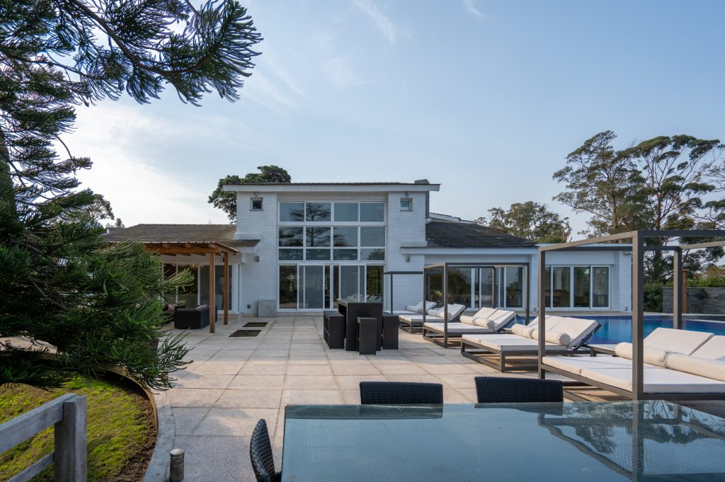 Área externa da casa em Punta del Este, no Uruguai. A casa possui dois andares, é branca, contém janelas grandes e portas de correr. Possui também uma piscina no lado direito da imagem.