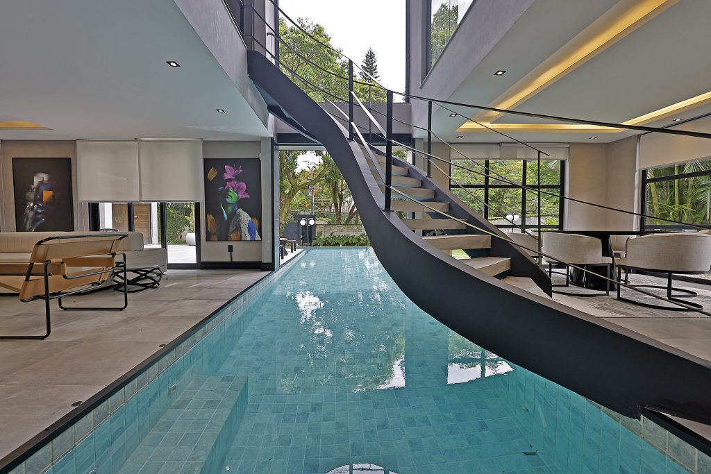 Imagem da arquitetura moderna da casa.
O espaço é divido pela piscina na vertical. Acima, no centro da foto e logo acima da piscina, tem a escada direto para o segundo andar.